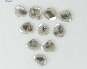 May include: Ten rough-cut, grey diamonds with irregular shapes are displayed. Each stone has a unique pattern of darker inclusions and a translucent appearance, set against a white background. The diamonds vary in size and shape.