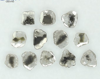 May include: A collection of rough-cut, irregular-shaped diamonds. The diamonds are primarily clear with dark gray and black inclusions. The image is taken on a white background. The logo "BHAVIN GEMS" is in the top left corner.