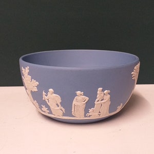 May include: A light blue ceramic bowl with a matte finish. The bowl features a white bas-relief design depicting classical figures and trees. The bowl is approximately 6 inches in diameter and 3 inches tall.