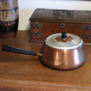 May include: A copper saucepan with a silver rim and lid, and a black handle. The pot is on a wooden surface. A wooden box and a decorative vase are in the background.