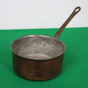 May include: A vintage copper saucepan with a long, brass handle. The pan has a rounded body and a slightly worn appearance, suggesting age and use. The handle is attached with a loop for hanging. The interior shows signs of use.