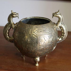 May include: Ornate brass-colored metal bowl with detailed relief carvings. The bowl has a round shape, three small feet, and two dragon-shaped handles. The relief depicts scenes of people and buildings. A decorative piece for home decor.