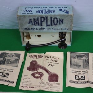 May include: Vintage Amplion pick-up and arm with volume control, displayed with its original box and promotional materials. The box is light blue with the Amplion logo and text. The arm is black.