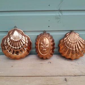 May include: Three copper-colored metal molds with hanging loops. Two molds are shaped like large scallop shells with ribbed textures, and the center mold is an oval shape featuring a raised fleur-de-lis design in the middle of the surface.