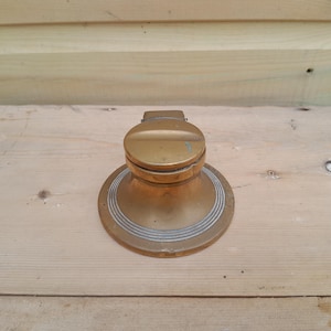 Op de afbeelding: Een vintage messing inktpot met een ronde basis en een scharnierend deksel. Het cirkelvormige ontwerp heeft concentrische ringen. Het messing heeft een aangetaste afwerking, wat duidt op ouderdom en gebruik. De inktpot is gemaakt van messing.