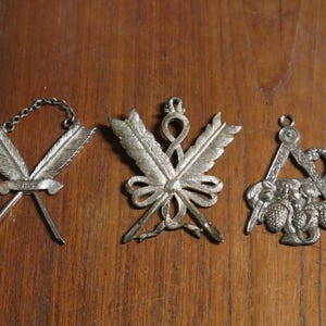 Puede incluir: Tres colgantes de metal plateado con diseños intrincados. Uno presenta plumas cruzadas y una pancarta, otro tiene plumas cruzadas con un lazo, y el tercero es un símbolo masónico con una brújula y un elemento decorativo.