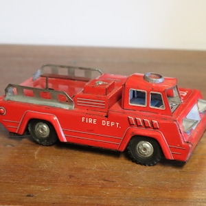 May include: A vintage red toy fire truck with the words "FIRE DEPT." printed on the side. The toy has black wheels, a silver ladder rack, and a silver siren on top. The toy is made of metal.