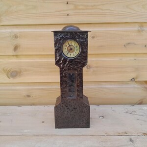 May include: A vintage brown metal clock with a textured finish. The clock has a round face with Roman numerals and ornate hands. The clock is rectangular with a small arched top and a base. The clock is approximately 30 cm tall.