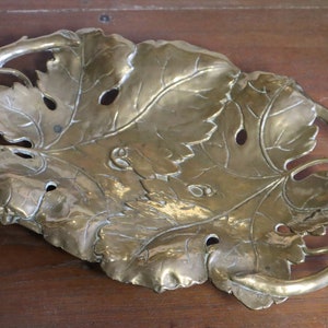 May include: A brass-colored decorative tray in the shape of a large leaf with intricate vein details and cut-out holes. The tray has a curved, branch-like handle and is likely made of metal. The overall design suggests a nature-inspired theme.