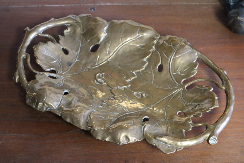 May include: A brass-colored decorative tray in the shape of a large leaf with intricate vein details and cut-out holes. The tray has a curved, branch-like handle and is likely made of metal. The overall design suggests a nature-inspired theme.