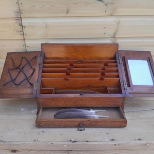May include: A vintage wooden writing desk, open to reveal storage compartments and a drawer with a feather. The desk has two side panels, one with a decorative crisscross design and the other with a mirror. The desk is made of dark wood and sits on a wooden surface.