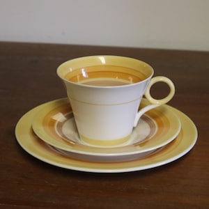 Op de afbeelding: Een witte theekop met een gele oor en een bijpassende schotel- en bordenset. De kop en borden hebben concentrische banden van geel, oranje en bruin. De set staat op een donkerbruin houten oppervlak.