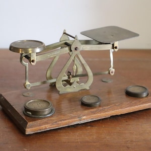 May include: An antique brass balance scale with a wooden base. The scale has two circular pans and a rectangular platform. Several brass weights are on the wooden base. The scale is likely used for measuring small items.