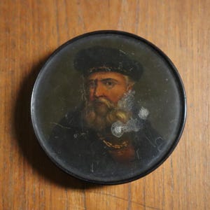 May include: A circular antique portrait miniature depicting a man with a beard and a dark hat. The portrait is set in a black metal frame, placed on a wooden surface. The painting has a vintage aesthetic.