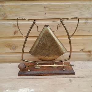 May include: A brass bell with a textured, triangular surface suspended within a curved brass frame. The frame is mounted on a rectangular wooden base, which also holds a thin brass rod with a round, dark-colored wooden mallet at one end.