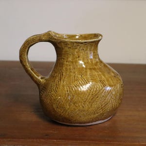 May include: A handmade ceramic jug with a rounded body and a curved handle. The jug has a warm, earthy brown colour with a glossy finish. The surface features a unique, carved pattern. The jug is approximately 15 cm tall.
