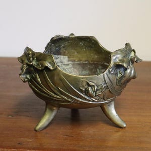 May include: An antique bronze bowl with a floral design. The bowl has three curved legs and is decorated with detailed leaf and flower motifs around the rim. The interior shows signs of age and use, with a patinaed finish.