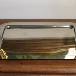 Puede incluir: Una bandeja rectangular plateada con esquinas redondeadas. La bandeja tiene asas con forma de piña en cada extremo. La bandeja es de metal brillante y refleja el entorno. Se encuentra sobre una superficie de madera.
