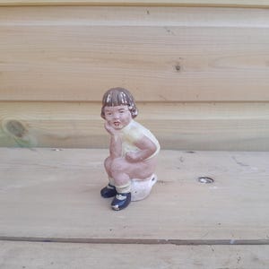 Op de afbeelding: Een vintage keramieken beeldje van een kind dat op een potje zit. Het beeldje is geschilderd in gedempte kleuren, met een lichtgeel shirt, bruin haar en zwarte schoenen. Het kind poseert met de kin op de hand.