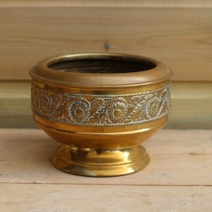 Könnte beinhalten: Vintage Messing-Blumentopf mit dekorativem Blumenmuster am Rand. Der Pflanztopf hat eine breite Öffnung und steht auf einem kleinen, runden Sockel. Das Messing hat einen warmen, goldenen Farbton.
