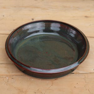 May include: A handmade ceramic pie dish with a dark green interior and a dark red rim. The dish is round and sits on a light brown wooden surface. The pie dish is glazed and has a glossy finish.