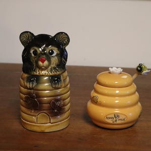 Könnte beinhalten: Zwei Honigtöpfe aus Keramik. Einer ist wie ein Bär geformt, der aus einem Bienenstock kommt, mit schwarzem Kopf und braunem Körper. Der andere ist ein gelber, bienenstockförmiger Topf mit einem bienenförmigen Löffel. Die Worte "honey de miel" stehen auf dem Topf.