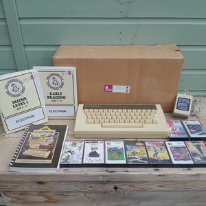 May include: An Acorn Electron computer with a beige keyboard, a power supply unit, a cardboard box, a spiral-bound user guide, two educational software binders titled Maths Level 1 and Early Reading, and eight assorted game cassettes in plastic cases.