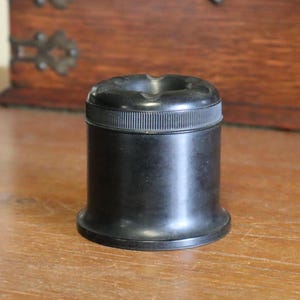 May include: A black, cylindrical ashtray with a ribbed band around the middle and a recessed top. The ashtray sits on a wooden surface, with a wooden cabinet in the background.