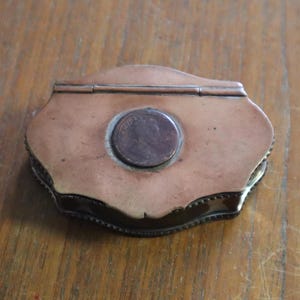 May include: An antique copper-coloured box with a hinged lid and a coin set into the top. The box has a decorative, scalloped edge and sits on a wooden surface.