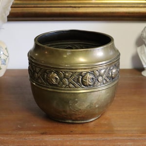 Könnte beinhalten: Ein Vintage-Pflanzgefäß aus Messing mit einem dekorativen Band aus geprägten Rosen und Blättern. Das Pflanzgefäß hat einen abgerundeten Körper und einen dunklen Innenraum. Es steht auf einer Holzoberfläche, mit einer Büste und einer Vase im Hintergrund.