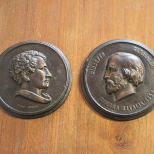 May include: Two antique bronze medallions with relief portraits. One depicts a man with curly hair, the other a bearded man. The round medallions have inscriptions. The background is a wooden surface.