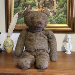 Può includere: Un orsacchiotto marrone vintage con naso cucito e occhi a bottone è seduto su una superficie di legno. L'orsacchiotto è circondato da oggetti decorativi, tra cui una statua in bronzo, un vaso con una piuma, un busto e un vaso di ceramica alto.