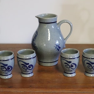 May include: A gray ceramic jug with a handle and four matching small cups. The jug and cups have blue decorative accents. The set is displayed on a wooden surface.