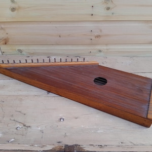 Puede incluir: Un instrumento musical de madera con forma trapezoidal. El instrumento tiene un acabado marrón oscuro con un agujero de sonido y varias clavijas de afinación. El instrumento está sobre una superficie de madera.
