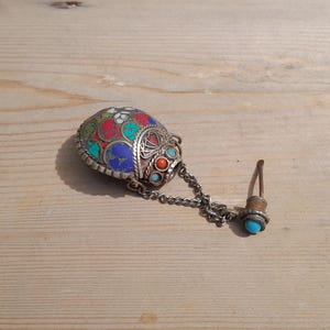 May include: A small, oval-shaped metal vessel featuring intricate wirework and inlaid colorful stone segments in shades of red, blue, green, and white. A silver-toned chain connects the main body to a matching stopper topped with a small blue stone.