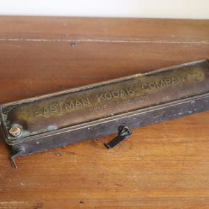 May include: Vintage Eastman Kodak Company developing box. Rectangular metal box with a brass-colored top and dark, aged sides. The text "EASTMAN KODAK COMPANY DEVELOPING BOX" is embossed on the top. The box has a small lever and a nozzle.