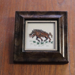 Peut inclure: Une œuvre d'art encadrée représentant une illustration de sanglier brun avec des défenses, sur fond blanc avec de l'herbe verte. L'œuvre est dans un cadre en bois brun foncé avec une bordure intérieure argentée.