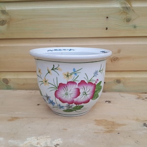 May include: A white ceramic plant pot with a rounded shape. The pot is decorated with pink and red flowers, green leaves, and small yellow and blue floral accents. A green line borders the top rim.