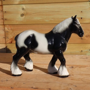 Pode incluir: Estatueta de cavalo de cerâmica em posição de pé. O cavalo é principalmente preto e branco, com cabeça e pernas pretas e corpo branco com manchas pretas. A crina e as penas são brancas. A estatueta está sobre uma superfície de madeira.