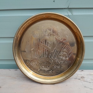 May include: A round brass tray featuring an etched illustration of a pixie dancing among tall grass, mushrooms, and a snail. The text "LUCKY PIXIE" is engraved on a small banner at the bottom. The tray has a polished metallic finish and a raised rim.