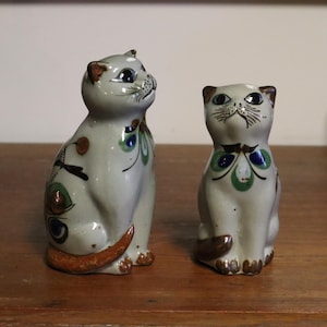 Peut inclure: Deux figurines de chat en céramique. Les chats sont gris clair avec des accents décoratifs marron, verts et bleus. L'un des chats regarde vers le haut, l'autre vers l'avant. Ils sont posés sur une surface en bois.