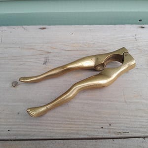 May include: A gold-toned brass nutcracker shaped like a pair of legs. The tool features two elongated handles that resemble human limbs, complete with detailed feet at the ends. The hinged mechanism is located at the top, designed for cracking nuts.