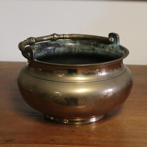 May include: A vintage brass cauldron with a handle. The pot has a rounded body and a dark interior. The handle is curved and attached to the sides of the pot. The cauldron is sitting on a wooden surface.
