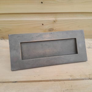 Pode incluir: Uma ranhura para cartas retangular de metal envelhecido com uma abertura rebaixada. A ranhura para cartas é de cor cinza suave e está colocada sobre uma superfície de madeira clara. O design é simples e funcional.