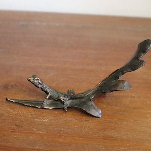 Op de afbeelding: Een bronzen sculptuur van een hagedis die rust op een gestileerd blad. De hagedis is gedetailleerd met een getextureerde huid en is geplaatst op een donkere, organisch gevormde basis. De sculptuur staat op een warme, houten ondergrond.