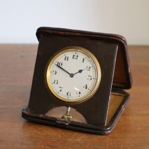 May include: A vintage brown leather travel clock with a white clock face and gold-coloured trim. The clock is in an open, rectangular case, and the clock face displays black numerals and hands. The clock is set on a wooden surface.