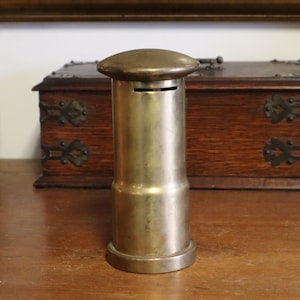 May include: A vintage brass money box with a slot at the top. The cylindrical bank has a rounded top and base. A wooden box and a decorative vase are in the background.