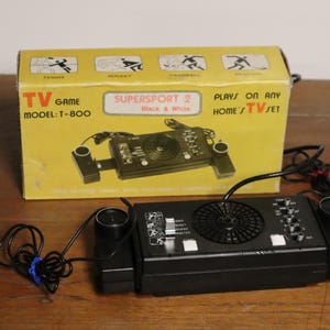 May include: A vintage black and white TV game console, model T-800, with a yellow box featuring sports icons. The console has two controllers and a central unit with dials and a speaker. The box reads "SUPERSPORT 2".