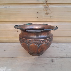 May include: A round copper planter with a wide, scalloped rim and a dark, weathered patina. The exterior features a band of hammered circular indentations above a series of embossed, petal-like shapes that extend toward the base of the vessel.
