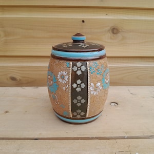 May include: A ceramic jar with a lid featuring a textured tan surface, a dark brown vertical band with white floral motifs, and bright turquoise accents. The jar has a barrel-like shape with horizontal turquoise stripes at the top and bottom.